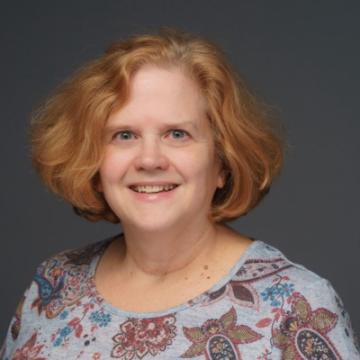 Photo of Susan Finkle-Sourlis. She's a white woman with red-ish hair in a bob cut. She's wearing a flower patterned shirt and in front of a dark gray background.